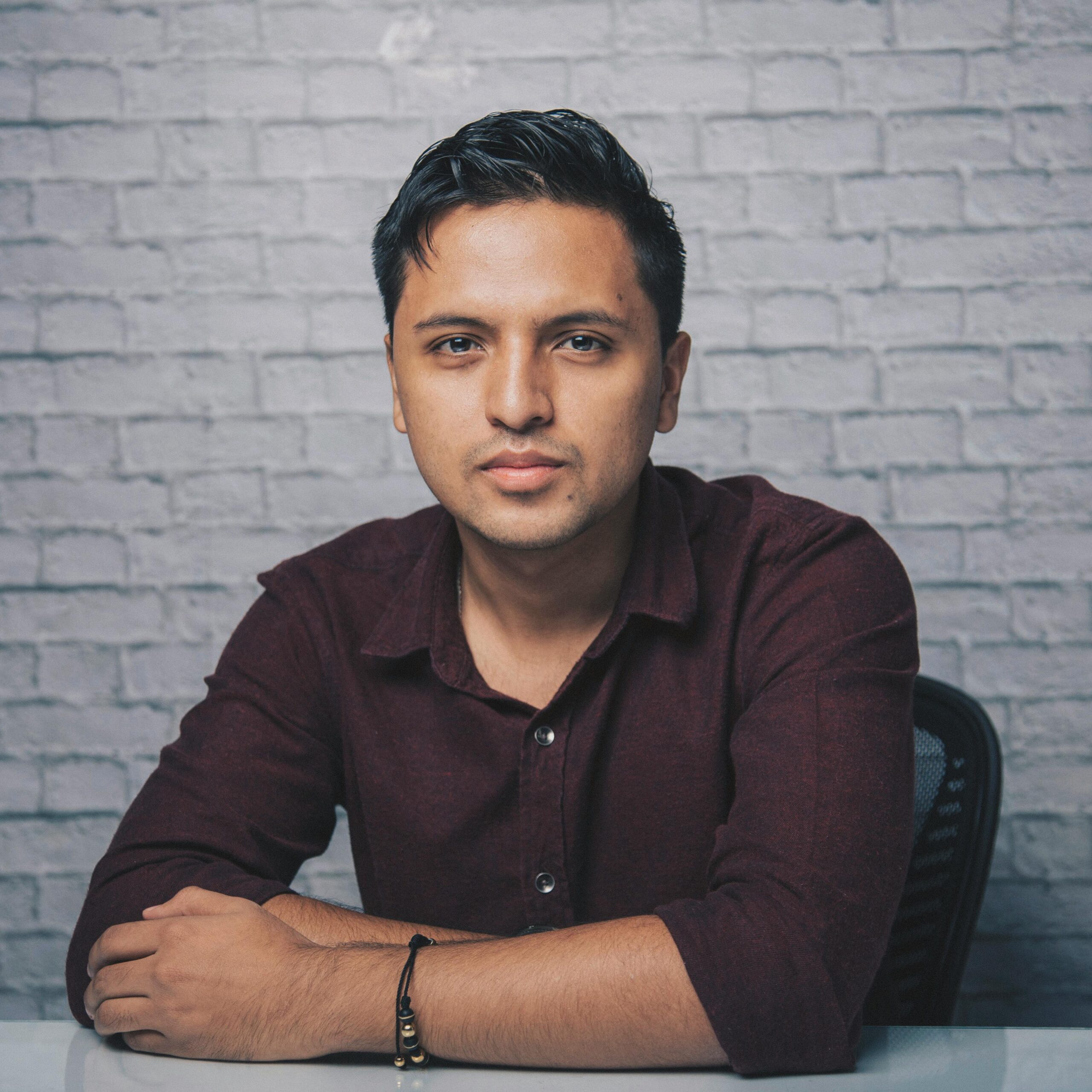 Confident adult Indian man in trendy shirt sitting at white table near brick wall while looking at camera in light room